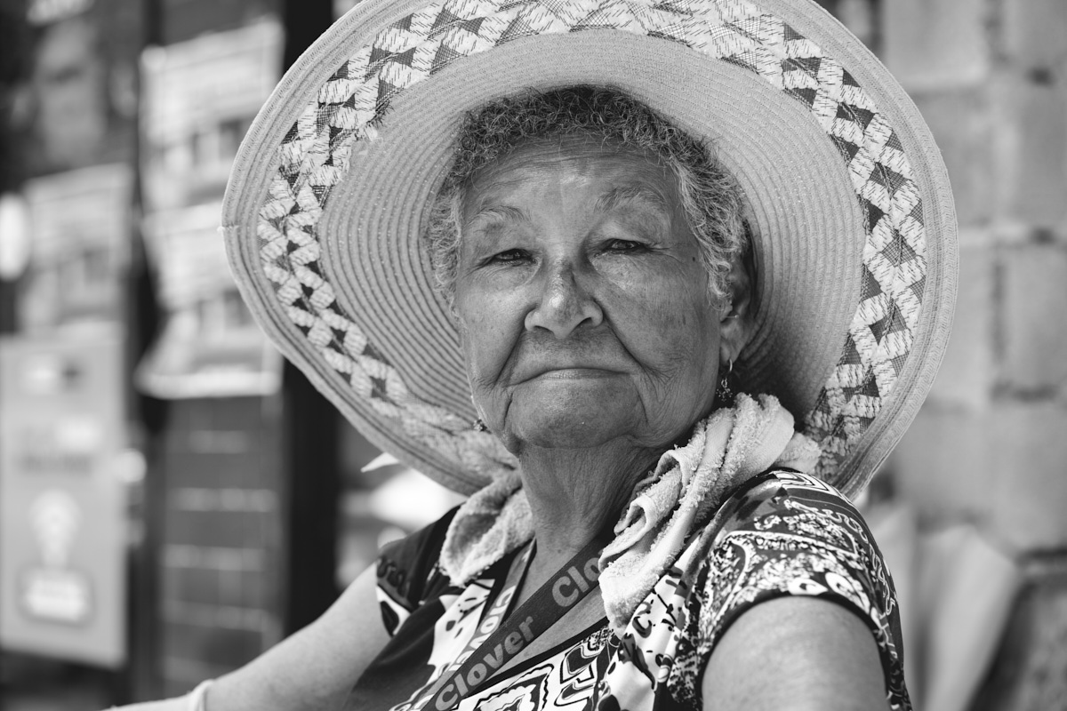 Black and white portrait of an elderly person wearing a decorative sun hat and patterned clothing.