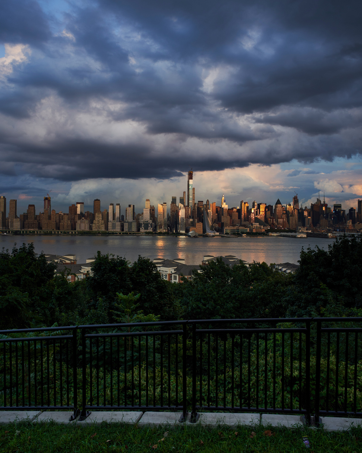 Dramatic cityscape of Manhattan skyline viewed from across the Hudson River, with lush green foreground and stormy clouds overhead. The dark clouds contrast with golden sunlight illuminating the skyscrapers.